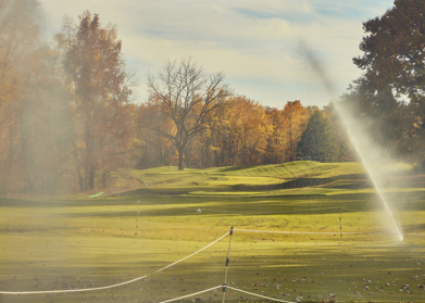 A golf course green with orange and brown trees behind it and sprinklers watering the ground.