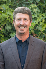 A portrait of a man with a dark dress shirt and a suit jacket.